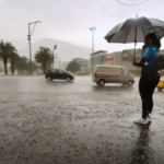 Persona con paraguas junto a una avenida de Quito durante una lluvia intensa, mientras varios vehículos circulan por la vía.