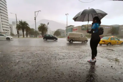 Persona con paraguas junto a una avenida de Quito durante una lluvia intensa, mientras varios vehículos circulan por la vía.